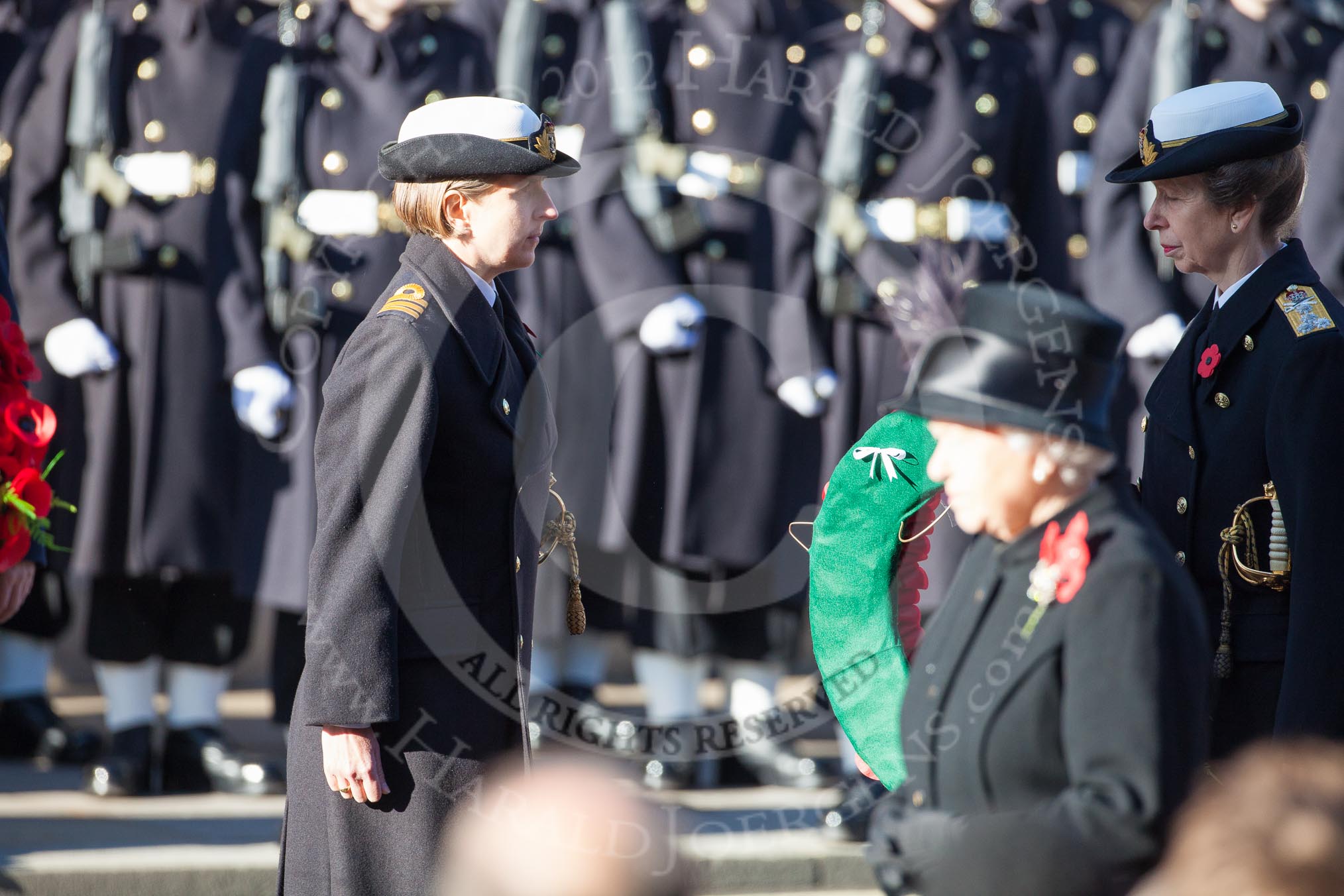 Commander Anne Sullivan, Royal Navy, handing, as Equerry, the wreath to HRH The Princess Royal. In the foreground, and out of focus, HM The Queen.